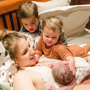 A woman lying in bed holding he rnew born baby is surrounded by her other children