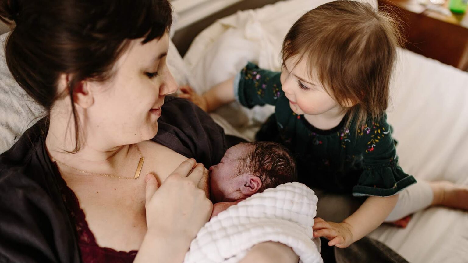 A woman nurses her baby while another child looks on